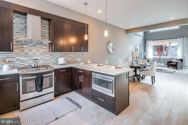 kitchen featuring pendant lighting, wall chimney range hood, light wood-type flooring, appliances with stainless steel finishes, and kitchen peninsula