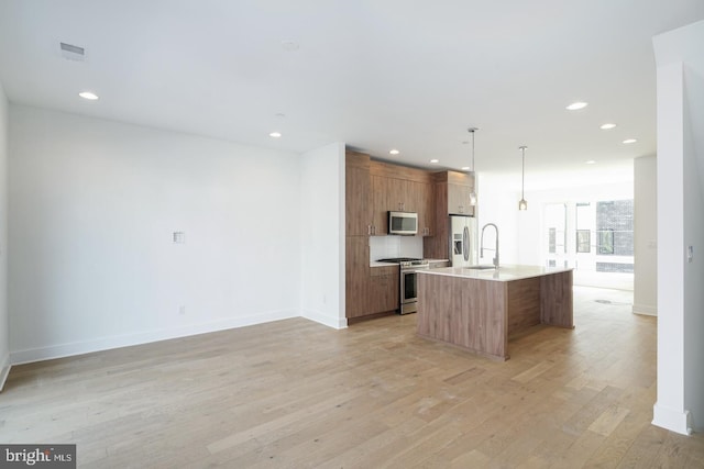 kitchen featuring a kitchen island with sink, sink, appliances with stainless steel finishes, decorative light fixtures, and light hardwood / wood-style floors