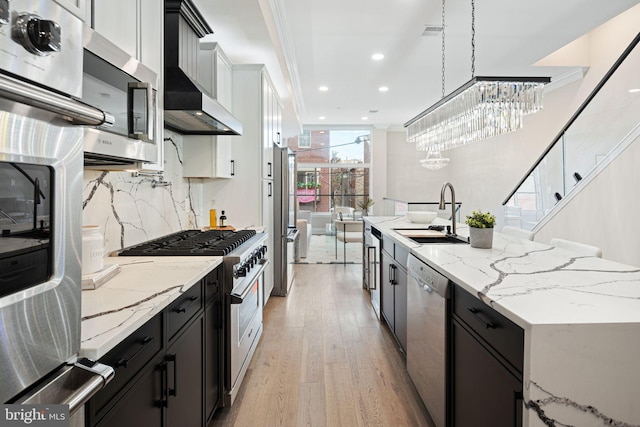 kitchen featuring light wood-type flooring, appliances with stainless steel finishes, decorative light fixtures, wall chimney range hood, and sink