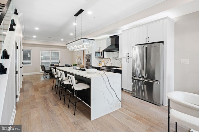 kitchen with stainless steel appliances, white cabinets, wall chimney exhaust hood, a kitchen island with sink, and light hardwood / wood-style flooring