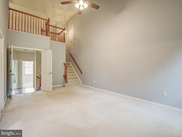 unfurnished living room featuring light carpet, a high ceiling, a ceiling fan, baseboards, and stairs