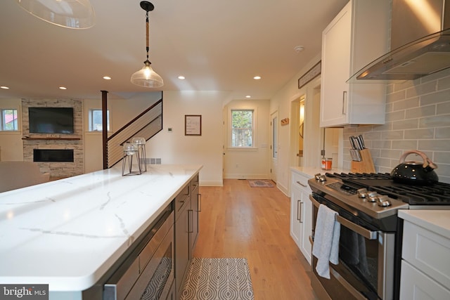 kitchen with light wood-type flooring, pendant lighting, stainless steel range with gas cooktop, white cabinets, and wall chimney exhaust hood