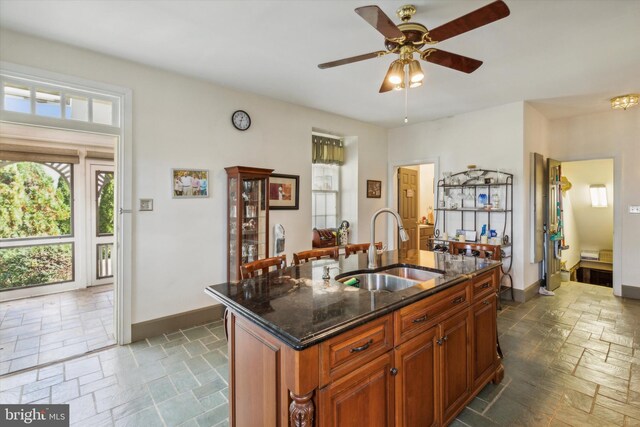 kitchen featuring a kitchen island with sink, sink, ceiling fan, and dark stone countertops