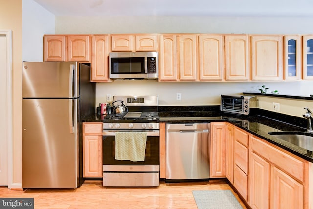 kitchen featuring stainless steel appliances, light brown cabinetry, sink, light hardwood / wood-style floors, and dark stone countertops