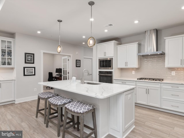 kitchen with white cabinets, sink, an island with sink, and wall chimney range hood