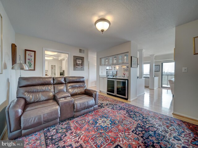 living room featuring light wood-type flooring, wine cooler, bar, ornate columns, and a textured ceiling