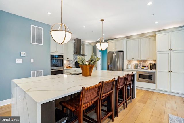 kitchen featuring light hardwood / wood-style floors, wall chimney range hood, pendant lighting, appliances with stainless steel finishes, and a large island with sink