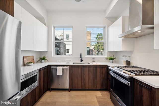 kitchen with light hardwood / wood-style floors, white cabinetry, sink, appliances with stainless steel finishes, and wall chimney exhaust hood