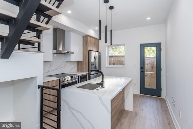 kitchen featuring stainless steel appliances, sink, wall chimney exhaust hood, decorative light fixtures, and a kitchen island with sink