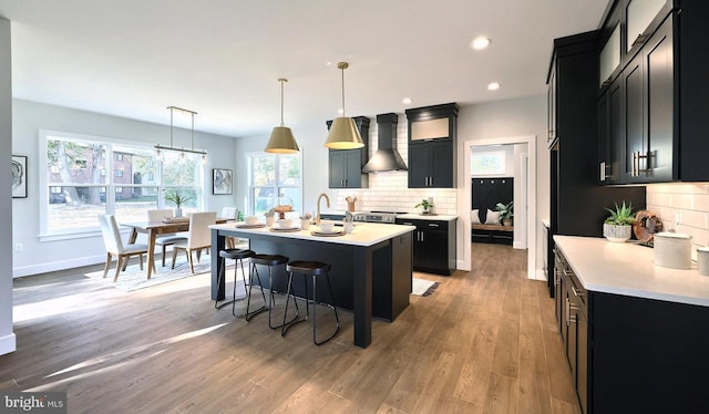 kitchen with wall chimney exhaust hood, dark cabinetry, decorative backsplash, an island with sink, and light countertops