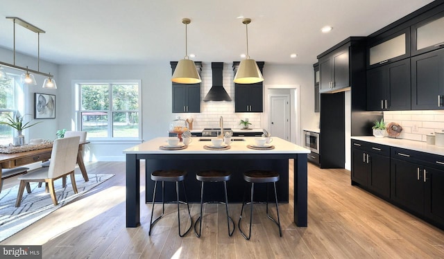 kitchen featuring light hardwood / wood-style flooring, tasteful backsplash, wall chimney range hood, and an island with sink