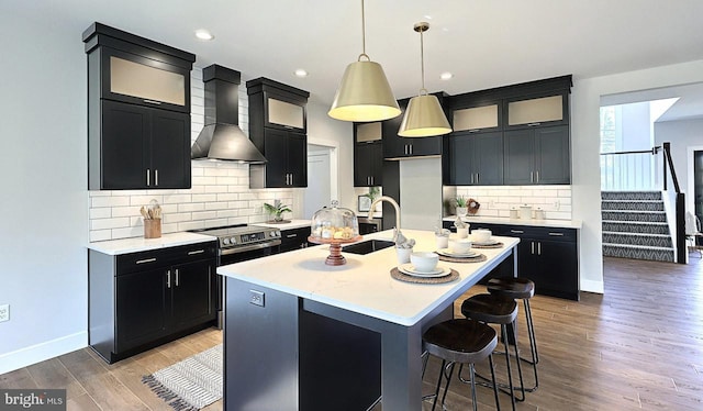 kitchen with wall chimney exhaust hood, decorative light fixtures, an island with sink, hardwood / wood-style floors, and decorative backsplash