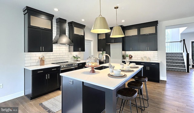 kitchen featuring wall chimney range hood, dark cabinetry, a sink, decorative backsplash, and light wood finished floors