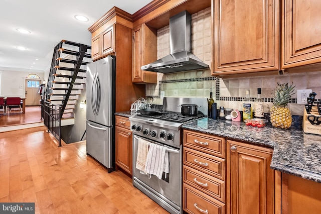 kitchen with stainless steel appliances, light hardwood / wood-style floors, tasteful backsplash, dark stone counters, and wall chimney range hood