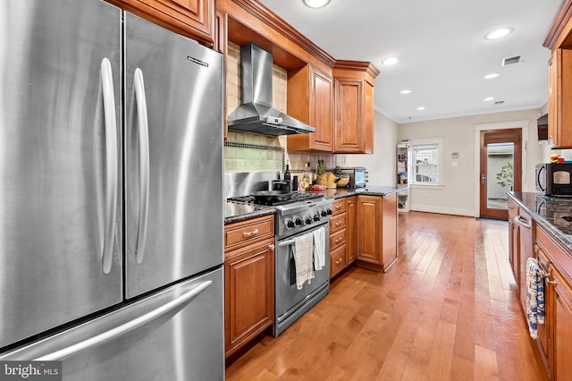 kitchen featuring stainless steel appliances, dark stone counters, wall chimney exhaust hood, tasteful backsplash, and light wood-type flooring