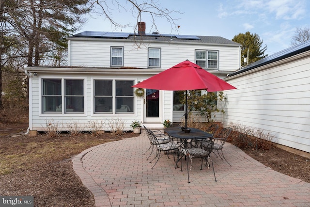 rear view of house featuring outdoor dining space, roof mounted solar panels, a chimney, and a patio