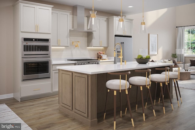 kitchen with wall chimney range hood, light wood-type flooring, an island with sink, appliances with stainless steel finishes, and white cabinetry