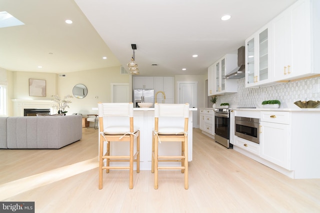 kitchen featuring stainless steel appliances, wall chimney range hood, decorative backsplash, white cabinets, and light hardwood / wood-style flooring