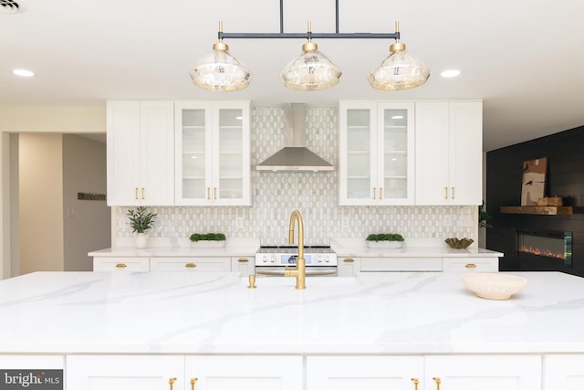 kitchen with decorative backsplash, wall chimney range hood, light stone countertops, white cabinetry, and decorative light fixtures