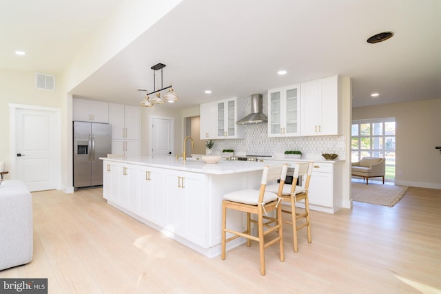 kitchen featuring stainless steel refrigerator with ice dispenser, wall chimney exhaust hood, a kitchen island with sink, white cabinetry, and light hardwood / wood-style flooring