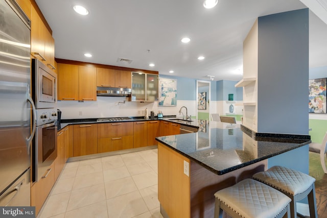 kitchen featuring kitchen peninsula, dark stone counters, built in appliances, a breakfast bar, and sink