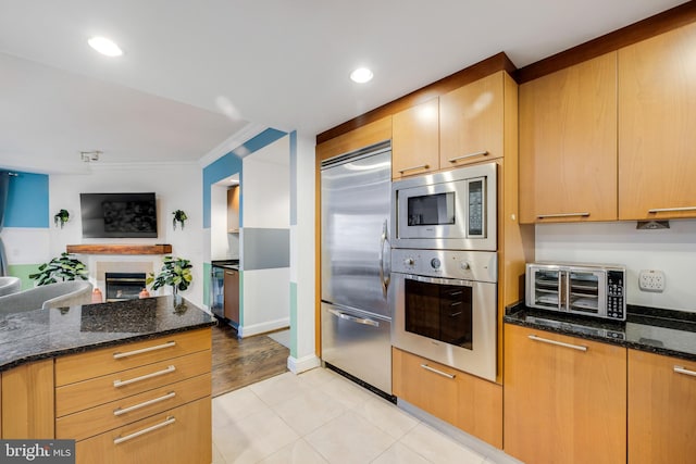 kitchen featuring light tile patterned flooring, ornamental molding, dark stone countertops, and built in appliances