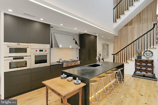 kitchen featuring stainless steel appliances, a spacious island, wall chimney range hood, sink, and light hardwood / wood-style flooring