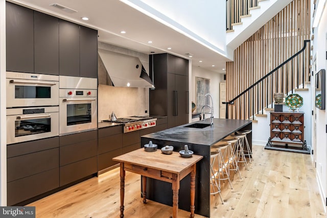 kitchen featuring wall chimney range hood, sink, light hardwood / wood-style flooring, an island with sink, and appliances with stainless steel finishes