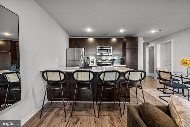 kitchen featuring dark brown cabinetry, a kitchen breakfast bar, light hardwood / wood-style flooring, decorative backsplash, and appliances with stainless steel finishes