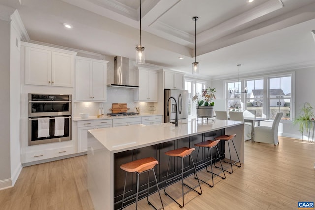 kitchen with appliances with stainless steel finishes, a large island with sink, pendant lighting, wall chimney exhaust hood, and light hardwood / wood-style flooring