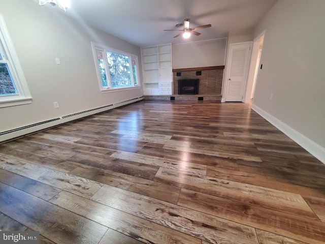 unfurnished living room featuring built in shelves, ceiling fan, dark wood-type flooring, a baseboard heating unit, and a fireplace