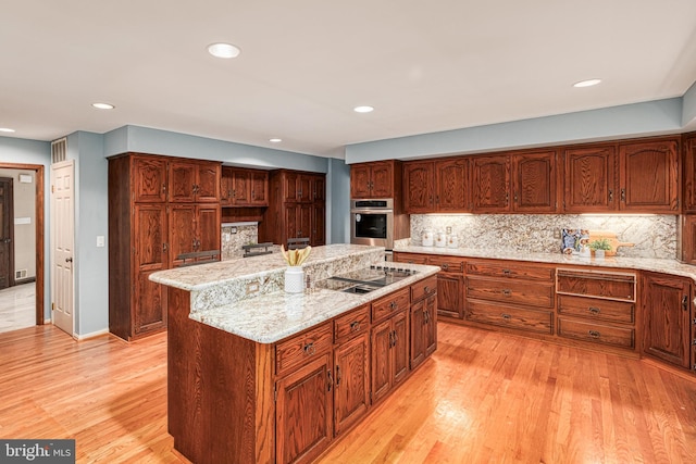 kitchen with tasteful backsplash, light stone counters, light wood-type flooring, an island with sink, and black electric cooktop