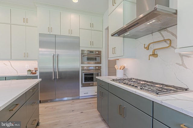 kitchen featuring appliances with stainless steel finishes, wall chimney range hood, light wood-type flooring, decorative backsplash, and gray cabinetry