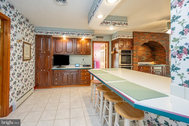 kitchen featuring double oven, light tile patterned floors, a breakfast bar, baseboard heating, and ceiling fan