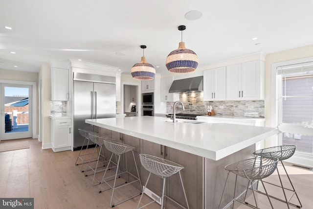 kitchen featuring wall chimney range hood, light hardwood / wood-style flooring, built in appliances, a large island, and white cabinetry