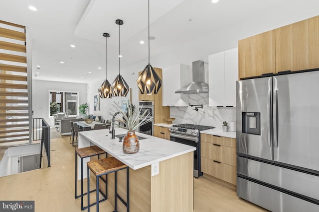 kitchen featuring stainless steel appliances, sink, wall chimney exhaust hood, an island with sink, and white cabinets