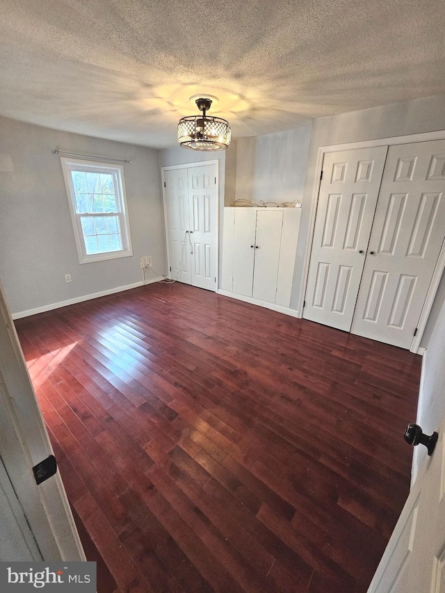 unfurnished bedroom with dark wood-style flooring, a chandelier, a textured ceiling, baseboards, and multiple closets