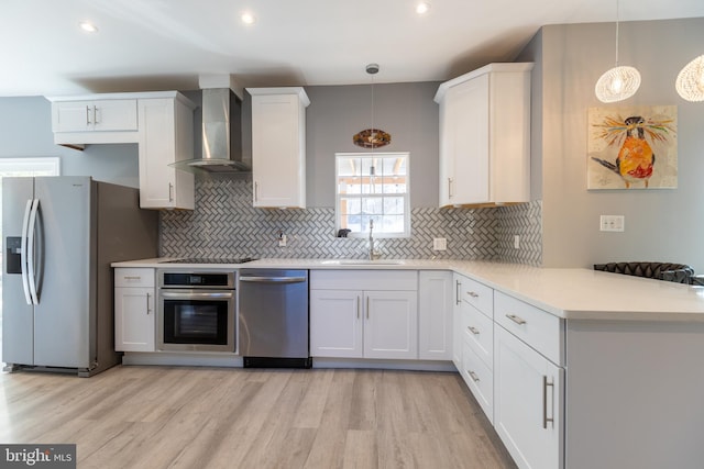 kitchen featuring stainless steel appliances, kitchen peninsula, wall chimney range hood, light wood-type flooring, and decorative light fixtures