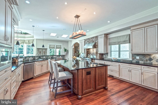 kitchen with wall chimney exhaust hood, stainless steel appliances, a kitchen island with sink, dark wood-type flooring, and decorative light fixtures