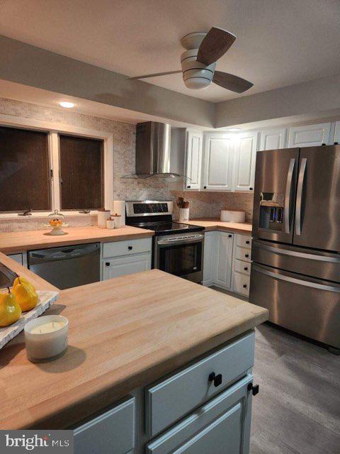 kitchen featuring butcher block countertops, wall chimney exhaust hood, white cabinetry, and appliances with stainless steel finishes