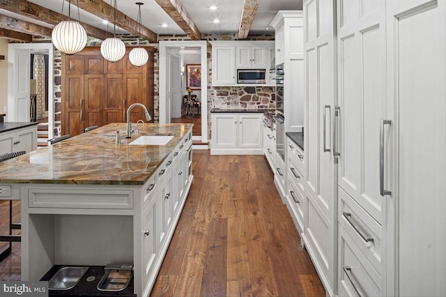 kitchen featuring dark stone counters, stainless steel microwave, hanging light fixtures, sink, and a kitchen island with sink