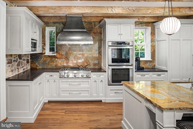 kitchen featuring stainless steel appliances, dark stone counters, decorative light fixtures, light wood-type flooring, and exhaust hood