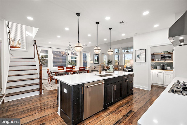 kitchen with a sink, light countertops, appliances with stainless steel finishes, wall chimney exhaust hood, and a center island with sink