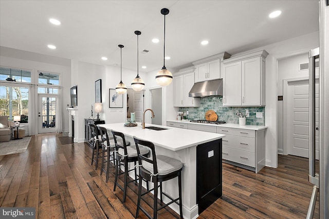 kitchen with under cabinet range hood, a kitchen island with sink, dark wood-style floors, and backsplash