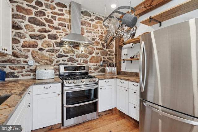 kitchen featuring white cabinetry, appliances with stainless steel finishes, wall chimney exhaust hood, dark stone countertops, and light hardwood / wood-style flooring