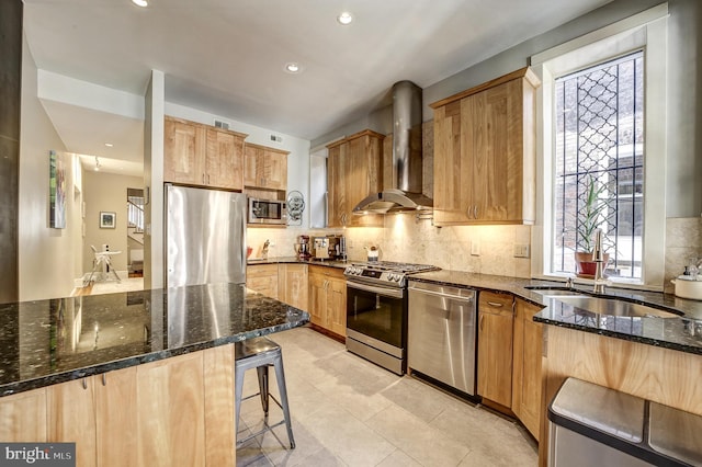 kitchen with a kitchen bar, stainless steel appliances, dark stone counters, sink, and wall chimney range hood