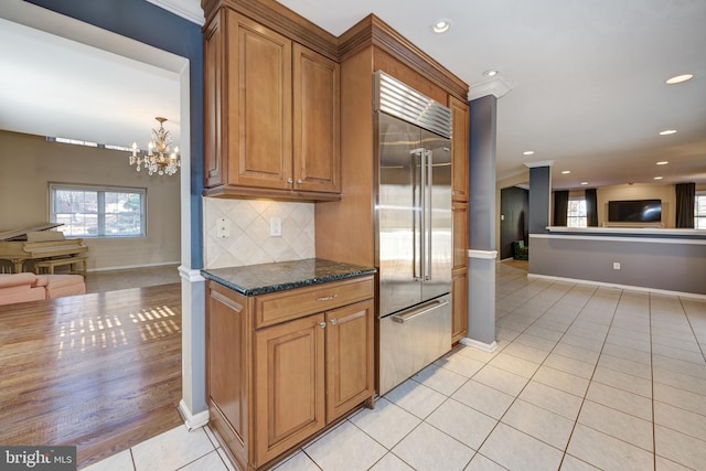 kitchen with tasteful backsplash, stainless steel built in refrigerator, a notable chandelier, light hardwood / wood-style floors, and dark stone counters