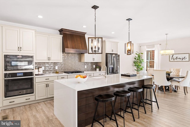 kitchen featuring a center island with sink, stainless steel refrigerator with ice dispenser, custom exhaust hood, light wood-type flooring, and pendant lighting