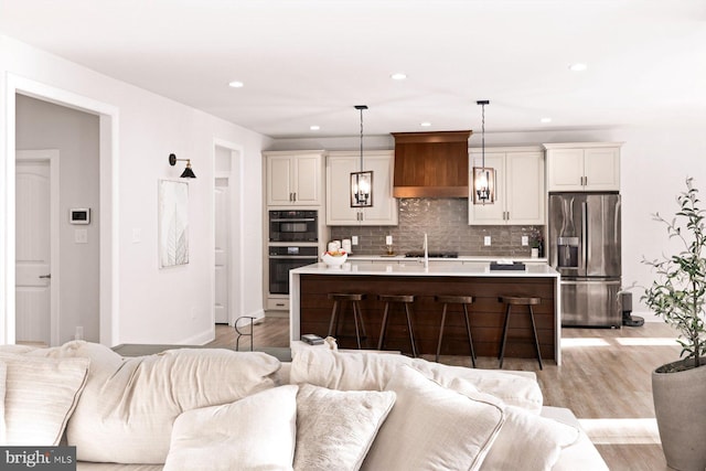 kitchen with stainless steel appliances, a center island with sink, custom exhaust hood, decorative light fixtures, and light hardwood / wood-style flooring