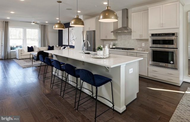 kitchen featuring a center island with sink, stainless steel appliances, dark hardwood / wood-style flooring, hanging light fixtures, and wall chimney exhaust hood